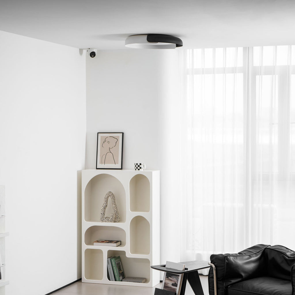 A white and black Clérachic ceiling light above a pale arched shelf, with a black leather armchair at the edge of the frame.