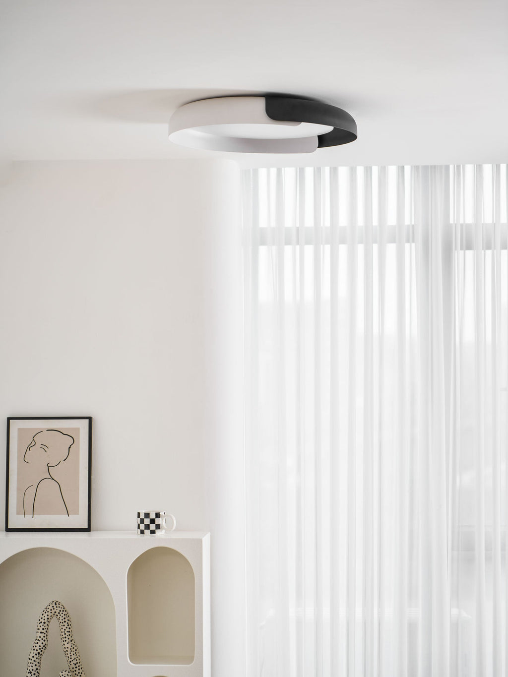 A black and white Clérachic ceiling light seen from below in a white room, with an arched shelf and a sheer curtain.