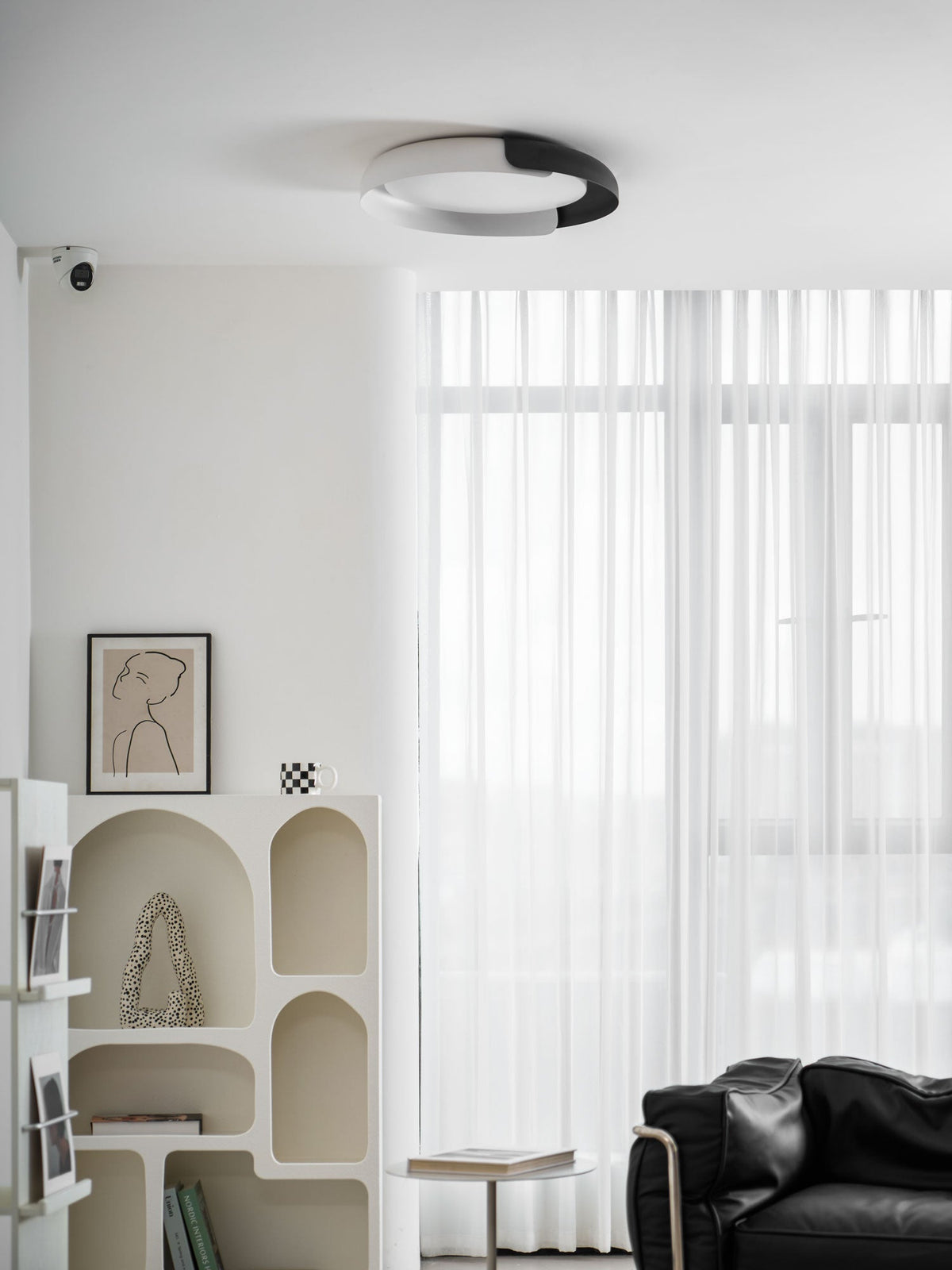 A black and white Clérachic ceiling light above a white arched shelf and sheer curtains, with a leather chair in the corner.
