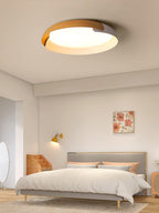 A light wood and white Clérachic ceiling light glows above a bed with peach cushions, in a pale bedroom with a brass wall light.