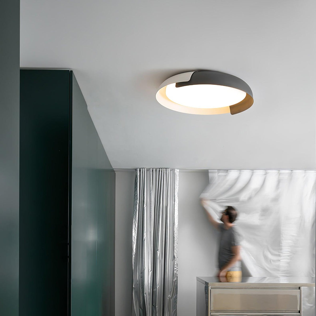 A white and black Clérachic ceiling light lit above a grey room with a dark green wall, where a person is hanging a sheer curtain.