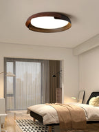 A dark wood and black Clérachic ceiling light lit over a bedroom with a tan throw, a floor lamp and a tall city-view window.