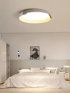 A white and light grey Clérachic ceiling light glows above a bed with white linen and a framed print, in a calm bedroom.