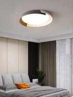 A grey and wood Clérachic ceiling light lit above a bed with an orange pillow, with brown curtains and a green plant.