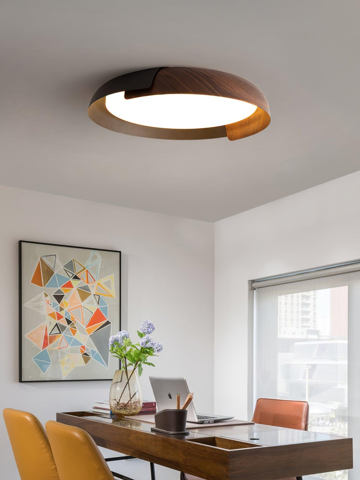 A black and dark wood Clérachic ceiling light lit above a desk with a laptop, orange chairs and a colourful geometric print.