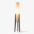 Slim Clérachic floor lamp with a tall white fabric cylinder shade on three dark walnut legs, lit and trailing a black cord on white.