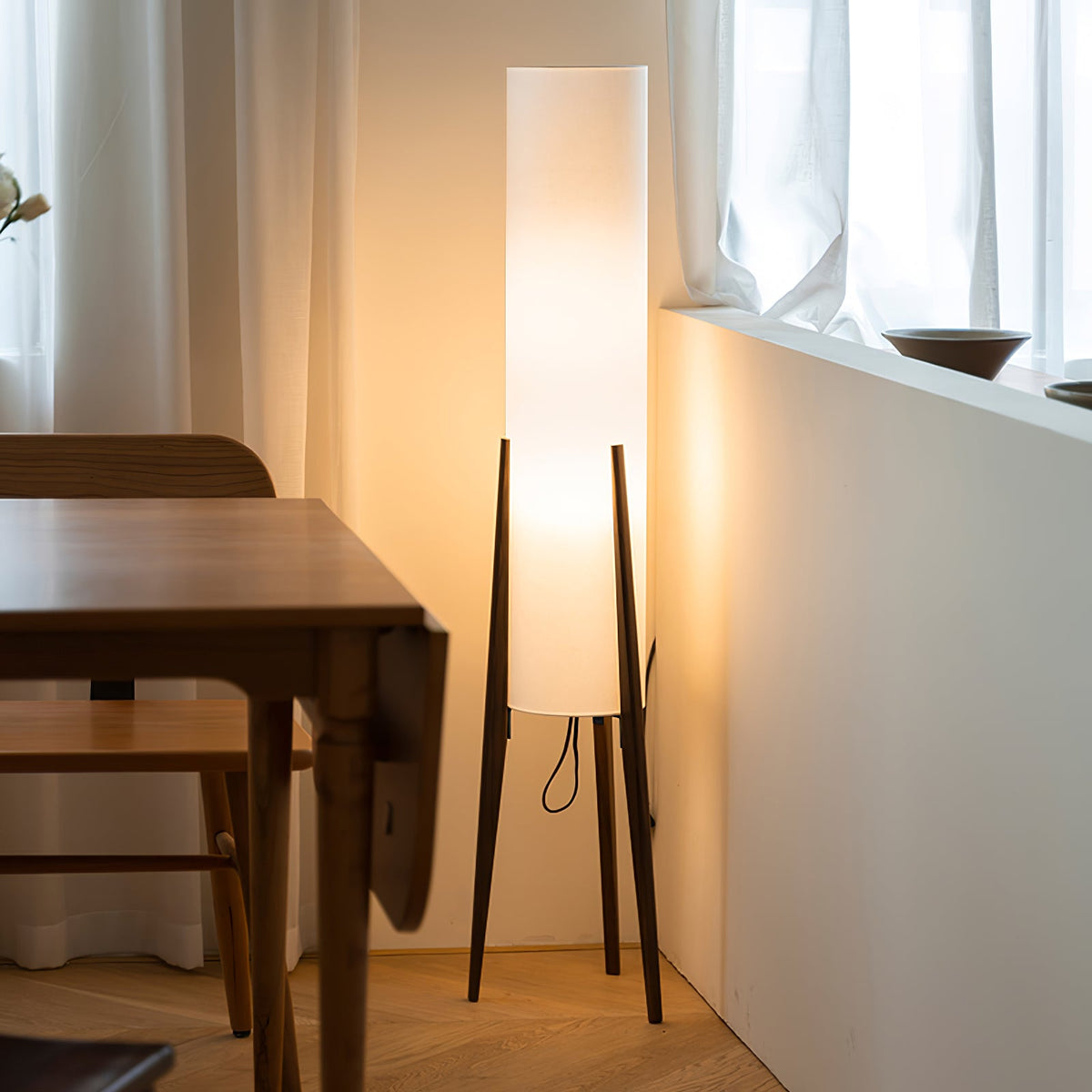 Lit Clérachic floor lamp with three walnut legs beside a wooden dining table, in a window corner with sheer curtains.
