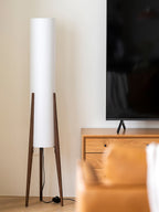Tall white cylinder shade on a Clérachic floor lamp with walnut tripod legs, standing by a wooden TV unit and a pale wall.