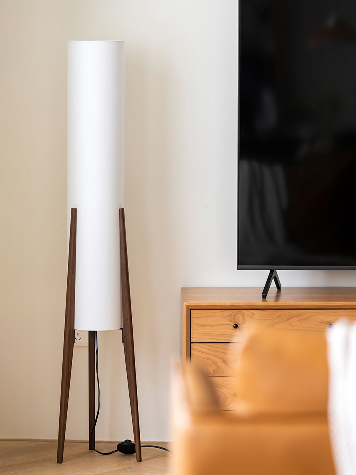 Tall white cylinder shade on a Clérachic floor lamp with walnut tripod legs, standing by a wooden TV unit and a pale wall.