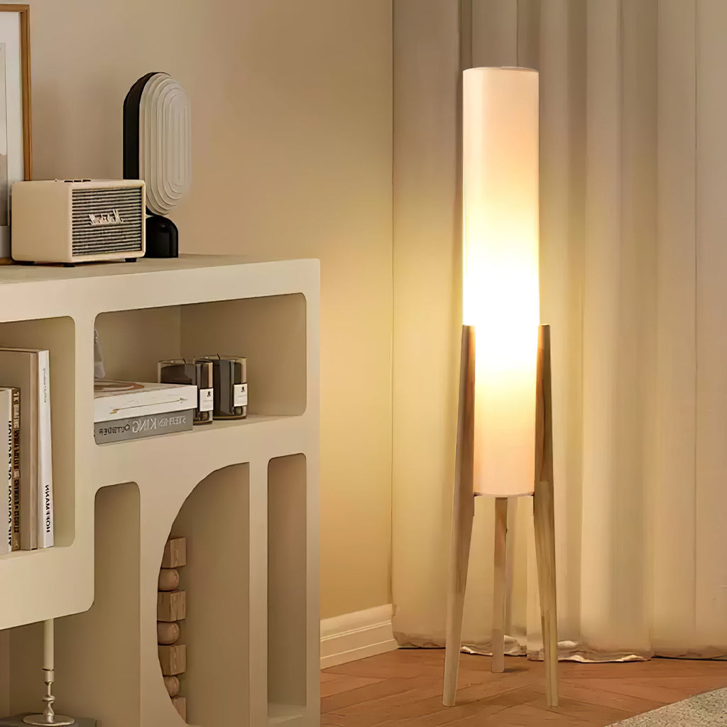 Clérachic floor lamp with a light wood tripod base glowing warm in a corner beside a cream shelf unit and linen curtains.