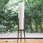 Walnut-legged Clérachic floor lamp with a white shade standing before a glass door onto green trees, with a foot switch on the cord.