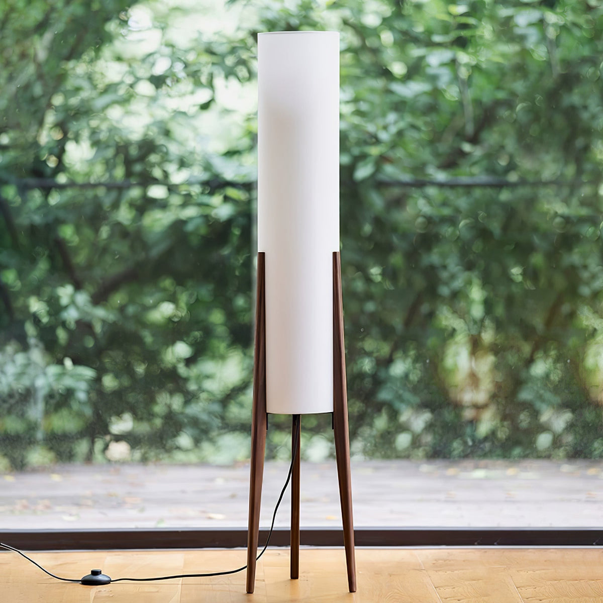 Walnut-legged Clérachic floor lamp with a white shade standing before a glass door onto green trees, with a foot switch on the cord.