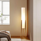 Lit Clérachic floor lamp on pale wood legs in a bedroom corner by a window, with a brown knit throw on the bed.