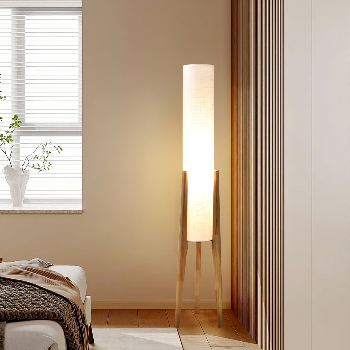 Lit Clérachic floor lamp on pale wood legs in a bedroom corner by a window, with a brown knit throw on the bed.