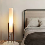 Clérachic floor lamp with a glowing white shade and walnut legs beside a bed with a rattan headboard and grey linen.