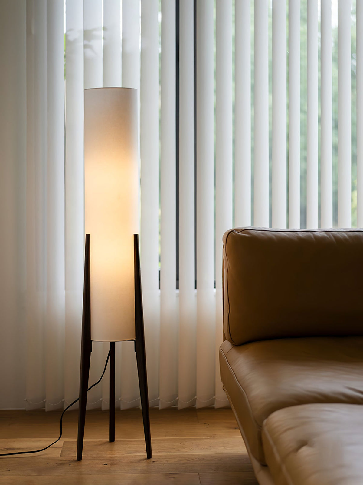Walnut tripod Clérachic floor lamp lit in front of white vertical blinds, next to a tan leather sofa.