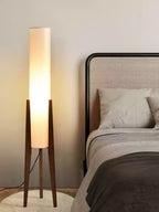 Lit slim Clérachic floor lamp on walnut legs, set on a round rug beside a bed with a rattan headboard and dark throw.