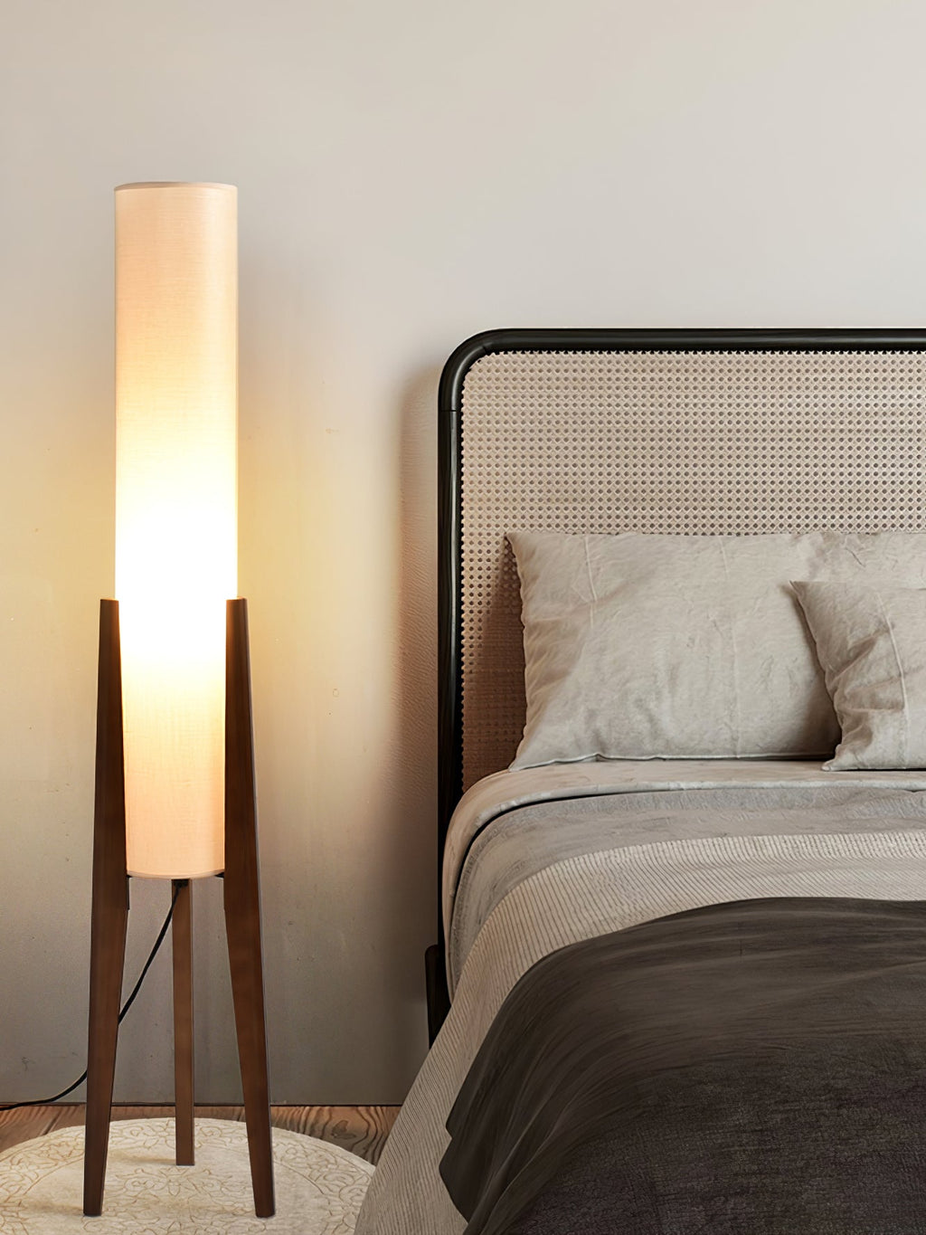 Clérachic floor lamp with a softly glowing white shade, seen beside a rattan-headed bed, its walnut legs on a round rug.