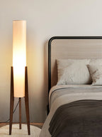Clérachic floor lamp with a softly glowing white shade, seen beside a rattan-headed bed, its walnut legs on a round rug.