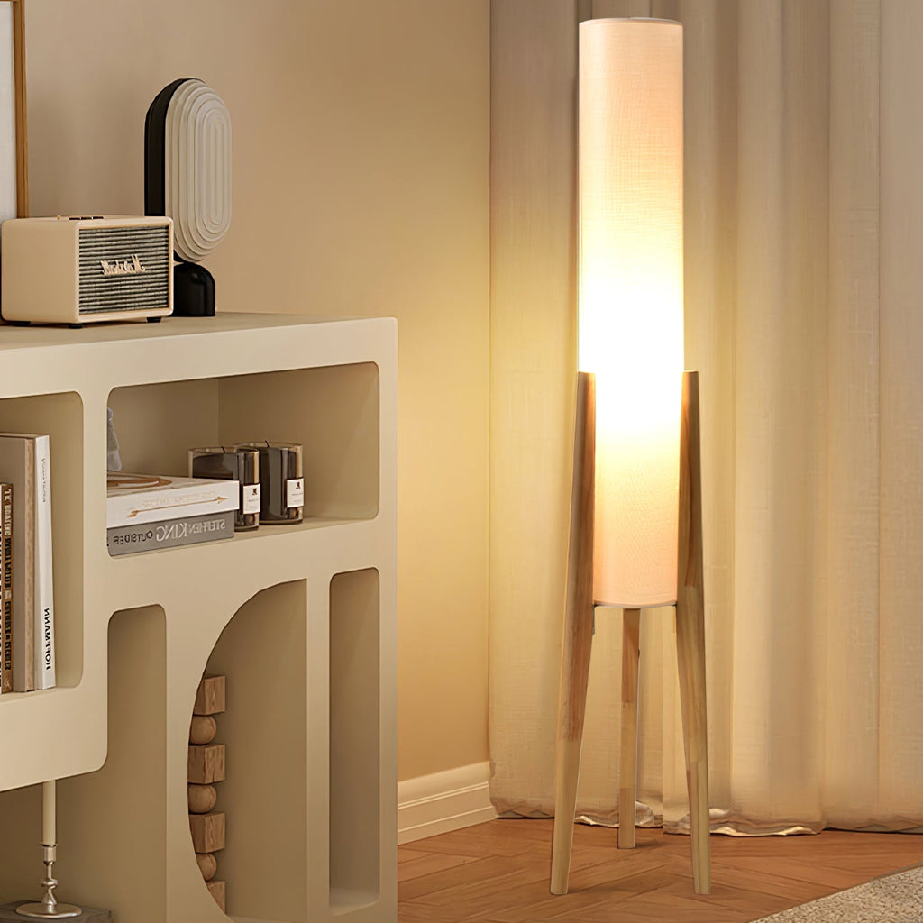Tall Clérachic floor lamp on light wood legs, lit, beside a cream shelf unit with books, a radio and linen curtains.