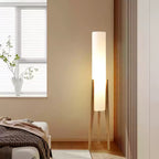 Light wood tripod Clérachic floor lamp lit in a bedroom corner, with a window and a vase of branches behind.