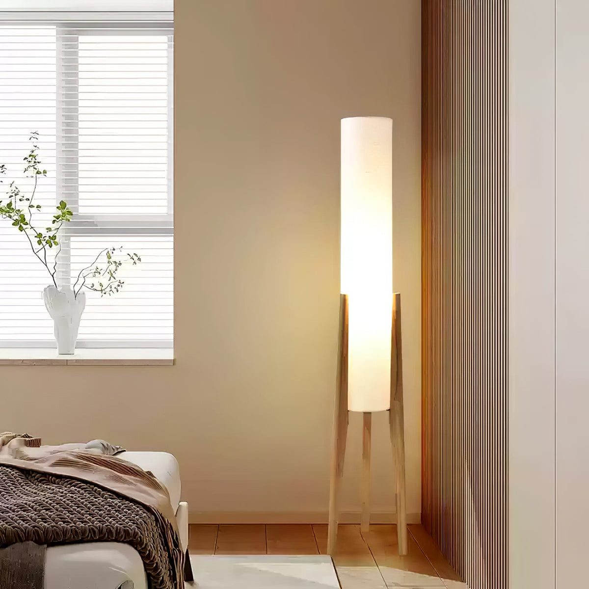 Light wood tripod Clérachic floor lamp lit in a bedroom corner, with a window and a vase of branches behind.