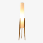 Clérachic floor lamp with a glowing white cylinder shade on three light wood legs, on a pale grey backdrop.