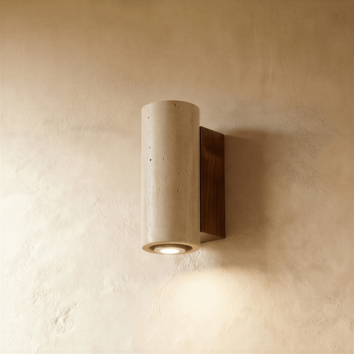 Clérachic wall lamp in a round pitted stone cylinder on a dark walnut mounting base, its downlight glowing below.