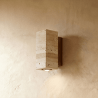 Clérachic wall lamp in rectangular pitted stone mounted on a dark walnut wood base, its downlight glowing on plaster.