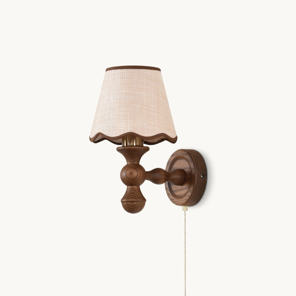 Clérachic wall light on a white backdrop, with a walnut-toned turned wood arm, a scalloped fabric shade and a pull chain.