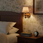 A Clérachic walnut wall light lit beside a dark wooden bed, with floral wallpaper, a stack of books and an alarm clock.