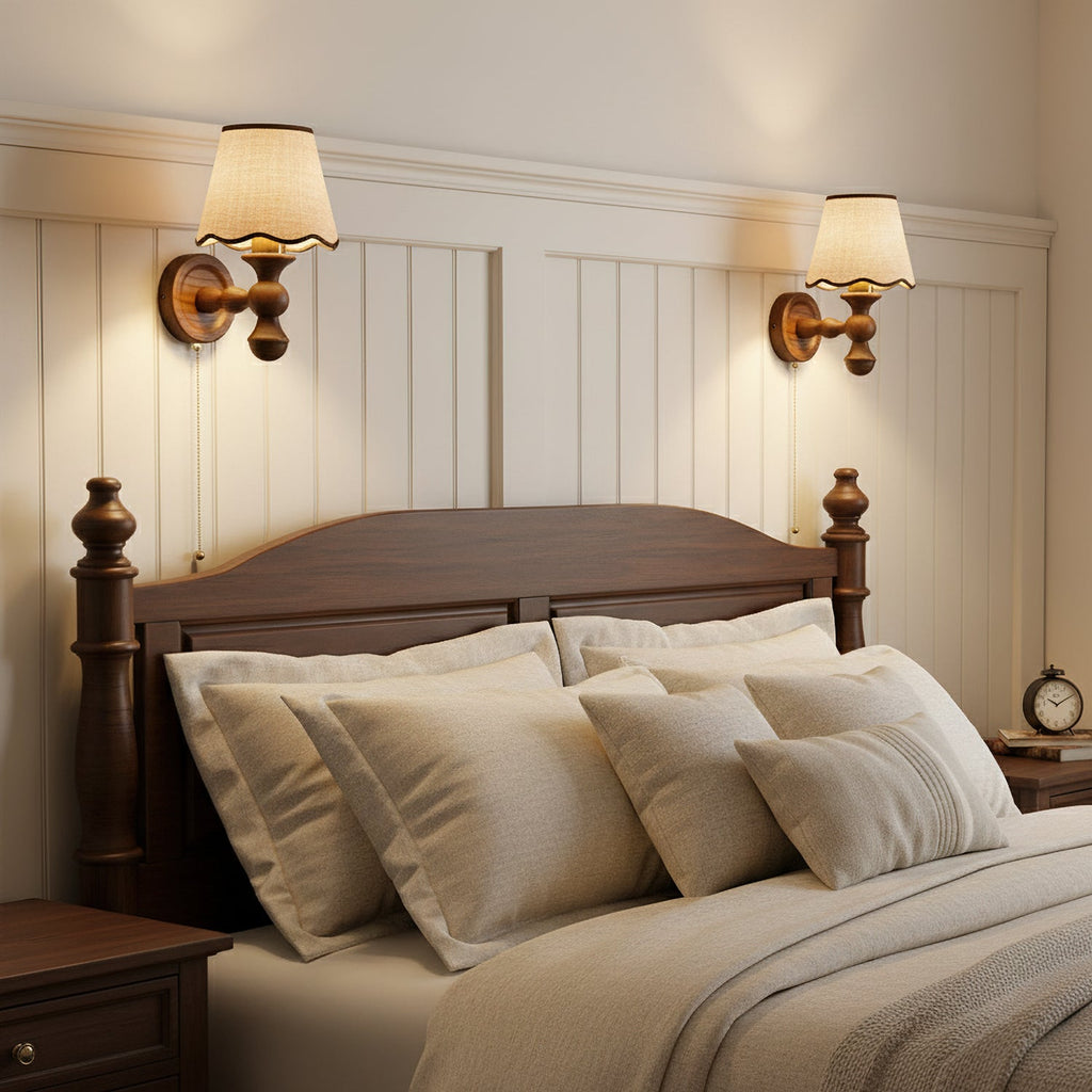 Two Clérachic wall lights lit above a bed with a dark wood headboard, on a white panelled wall with layered cushions.
