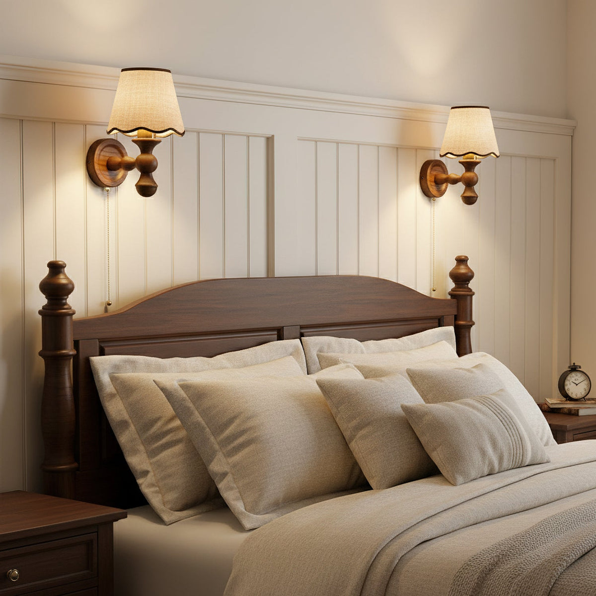 Two Clérachic wall lights lit above a bed with a dark wood headboard, on a white panelled wall with layered cushions.