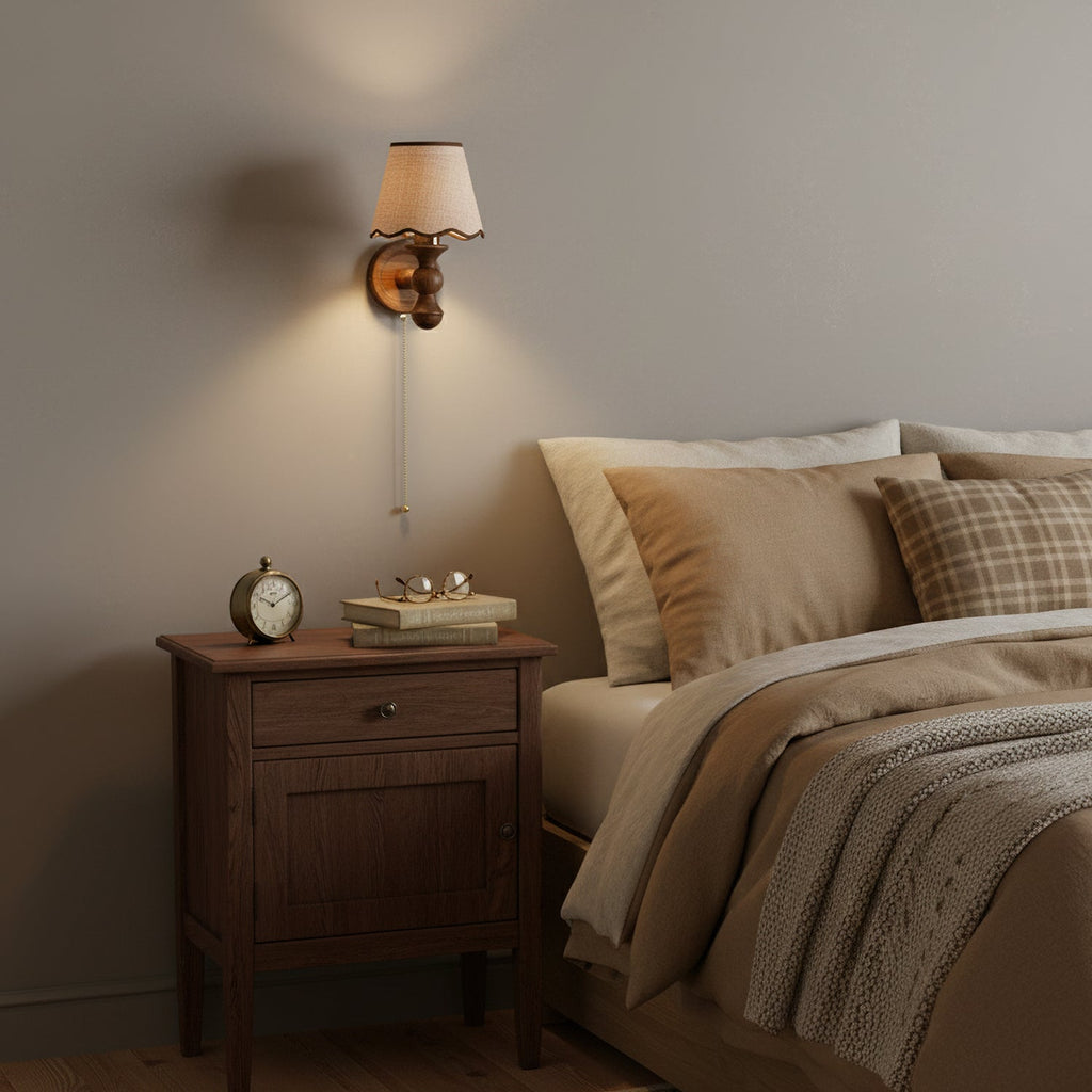 Clérachic walnut wall light lit above a wooden bedside cabinet with an alarm clock, books and glasses on top.