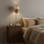 Clérachic walnut wall light lit above a wooden bedside cabinet with an alarm clock, books and glasses on top.