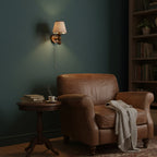 A Clérachic wall light lit on a dark green wall above a brown leather armchair and a small round table.