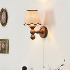 Close view of this Clérachic wall light on a white wall, its scalloped shade lit and a pull chain hanging down.