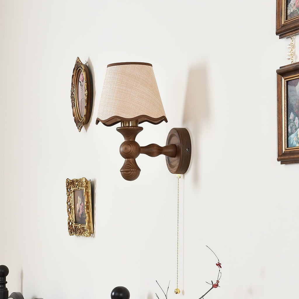 A Clérachic wall light on a white wall beside small gilt-framed pictures, with its walnut arm and pull chain visible.