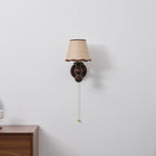 Clérachic wall light on a pale grey wall, its pull chain with a brass bead hanging above a wooden drawer unit.