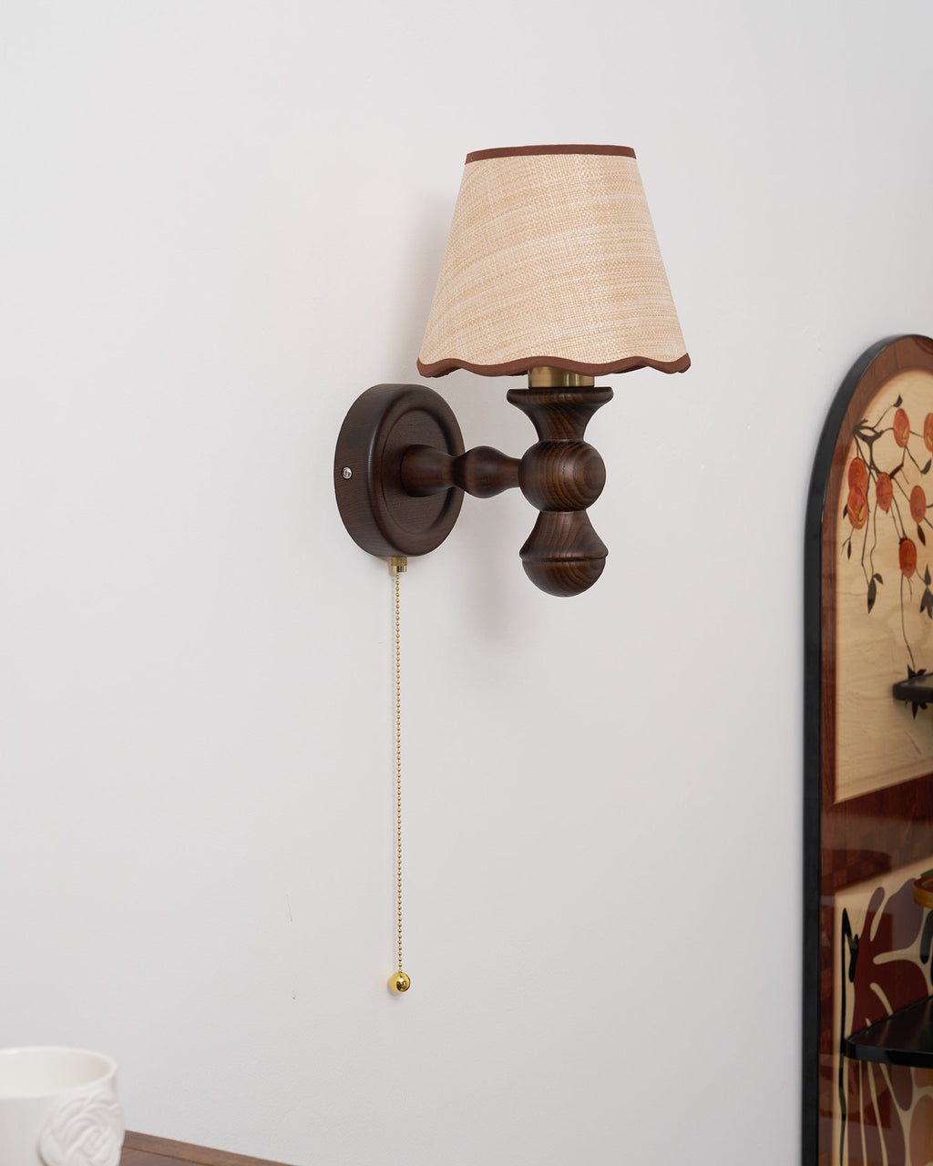 Side-on view of this Clérachic walnut wall light, with its round backplate, turned arm and scalloped shade.