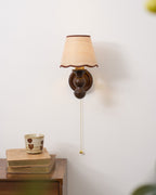A Clérachic wall light on a pale wall above a stack of books with a patterned cup on a wooden cabinet.