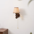 A Clérachic wall light on a white wall, its pull chain hanging beside a wooden cabinet with a pattern-printed cup.