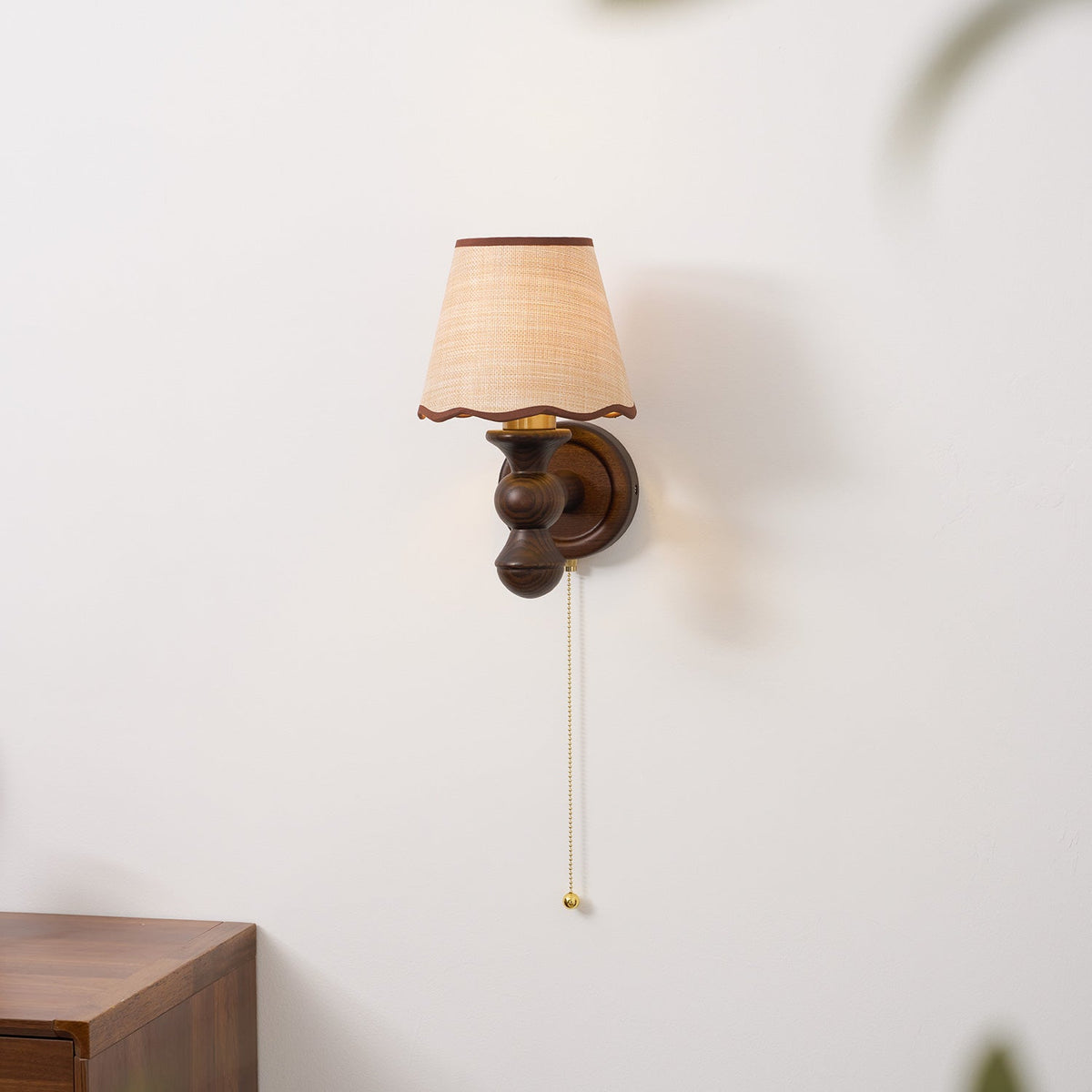 A Clérachic wall light on a white wall, its pull chain hanging beside a wooden cabinet with a pattern-printed cup.