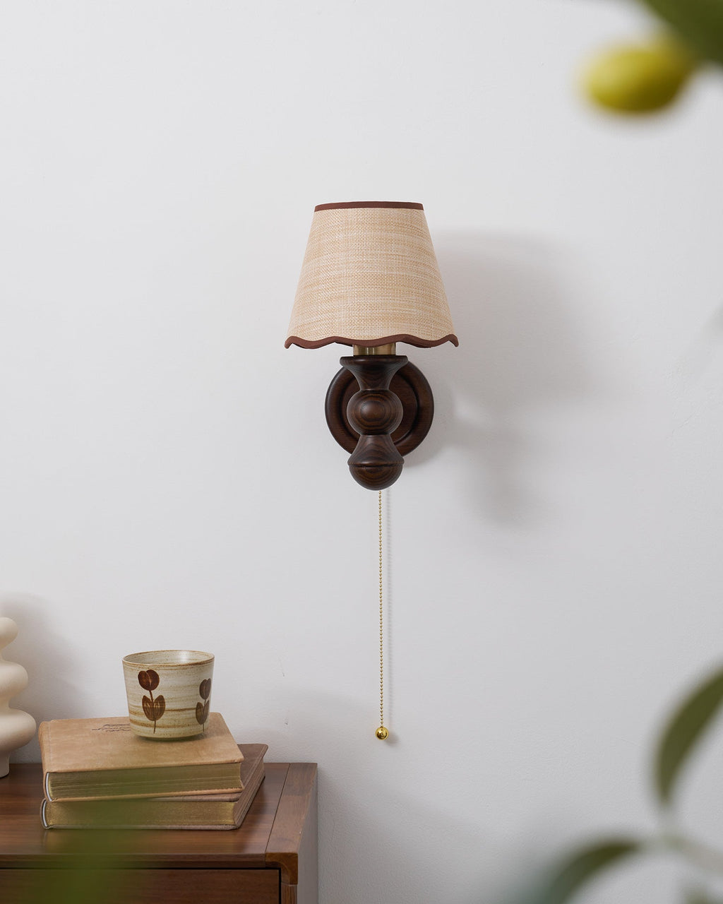 A Clérachic wall light on a pale wall above a cabinet with books and a cup, a leaf blurred at the edge.