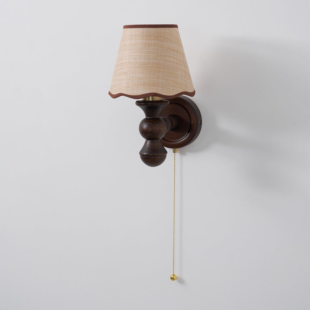 A Clérachic walnut wall light on a pale grey wall, shown from a slight angle with a long pull chain.