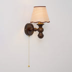 A Clérachic walnut wall light on a pale grey wall with a pull chain hanging down, the shade glowing softly.