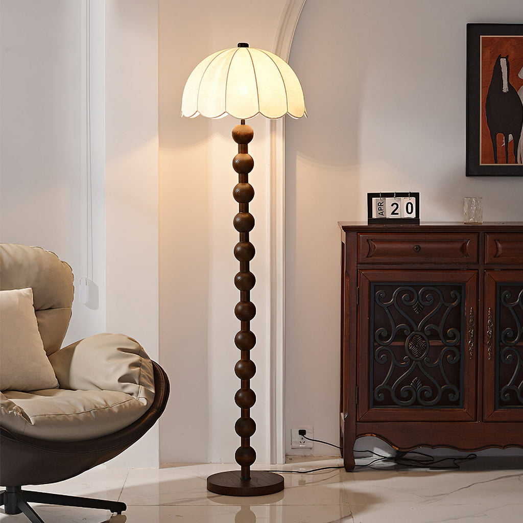 Lit dome shade above a stem of stacked wooden balls, beside a carved dark wood cabinet and a lounge chair; a Clérachic floor lamp.