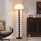 Lit dome shade above a stem of stacked wooden balls, beside a carved dark wood cabinet and a lounge chair; a Clérachic floor lamp.