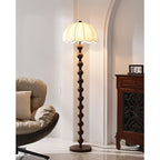 The lit scalloped shade sits in a bright corner with a plush chair and a dark cabinet, near an arch; a Clérachic floor lamp.