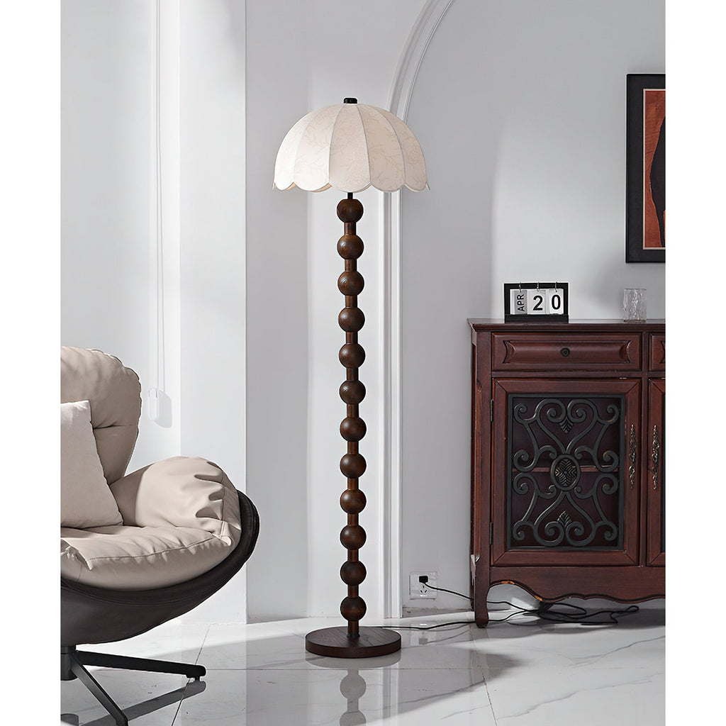 The unlit beige shade and the walnut-toned ball stem stand against a pale wall, with a cabinet and a chair; this Clérachic floor lamp.